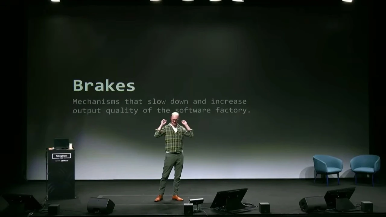 A presenter stands before a slide defining brakes as mechanisms that slow down and increase software factory output quality.